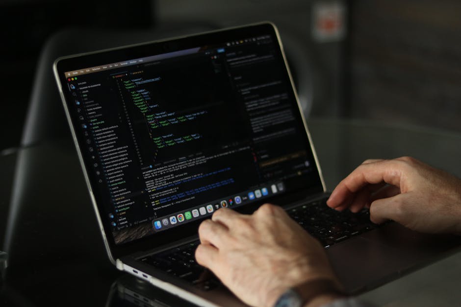 A close-up of a person's hands working on a laptop with a dark screen displaying lines of colorful code, placed on a black reflective surface. The background is blurred, showing a cozy indoor setting 