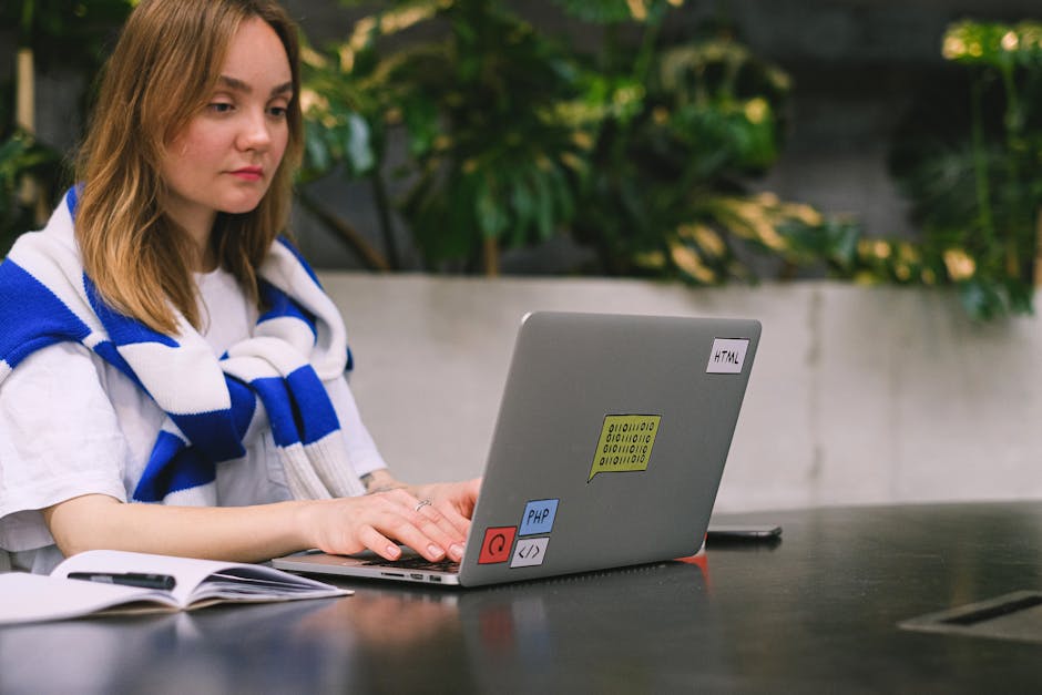 A young woman with shoulder-length blonde hair and wearing a white shirt, has a blue and white striped scarf draped over her shoulders. She is seated outdoors at a dark wooden table, working on a silv